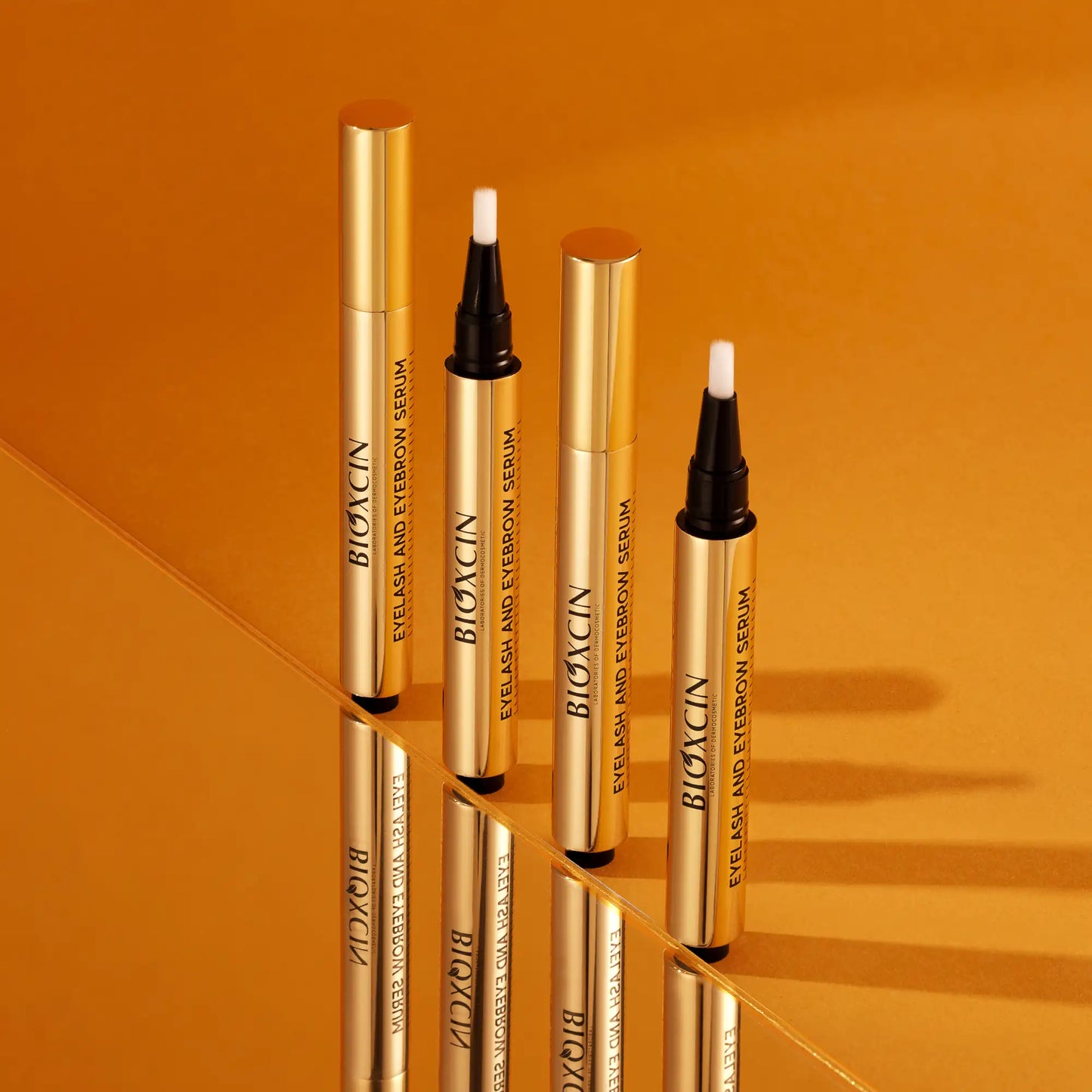 Two Bioxcin eyelash and eyebrow serum bottles with applicators on an orange background