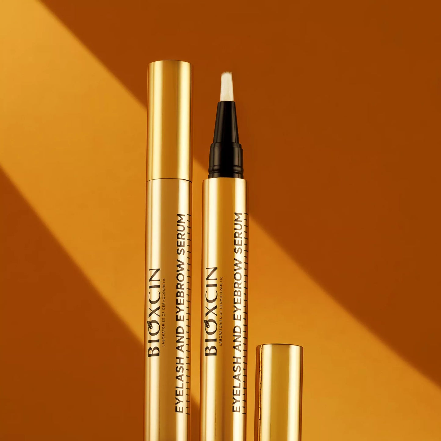 Two gold BIOXCIN eyelash and eyebrow serum bottles on an orange background