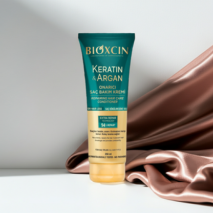 BIOXCIN Keratin & Argan hair care cream in a teal and gold squeeze tube, styled on a white surface with a draped rose gold satin fabric in the background.
