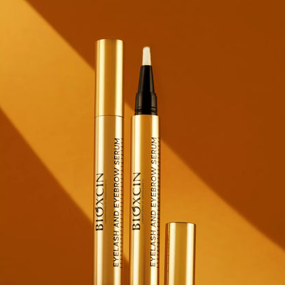 Two gold BIOXCIN eyelash and eyebrow serum bottles on an orange background