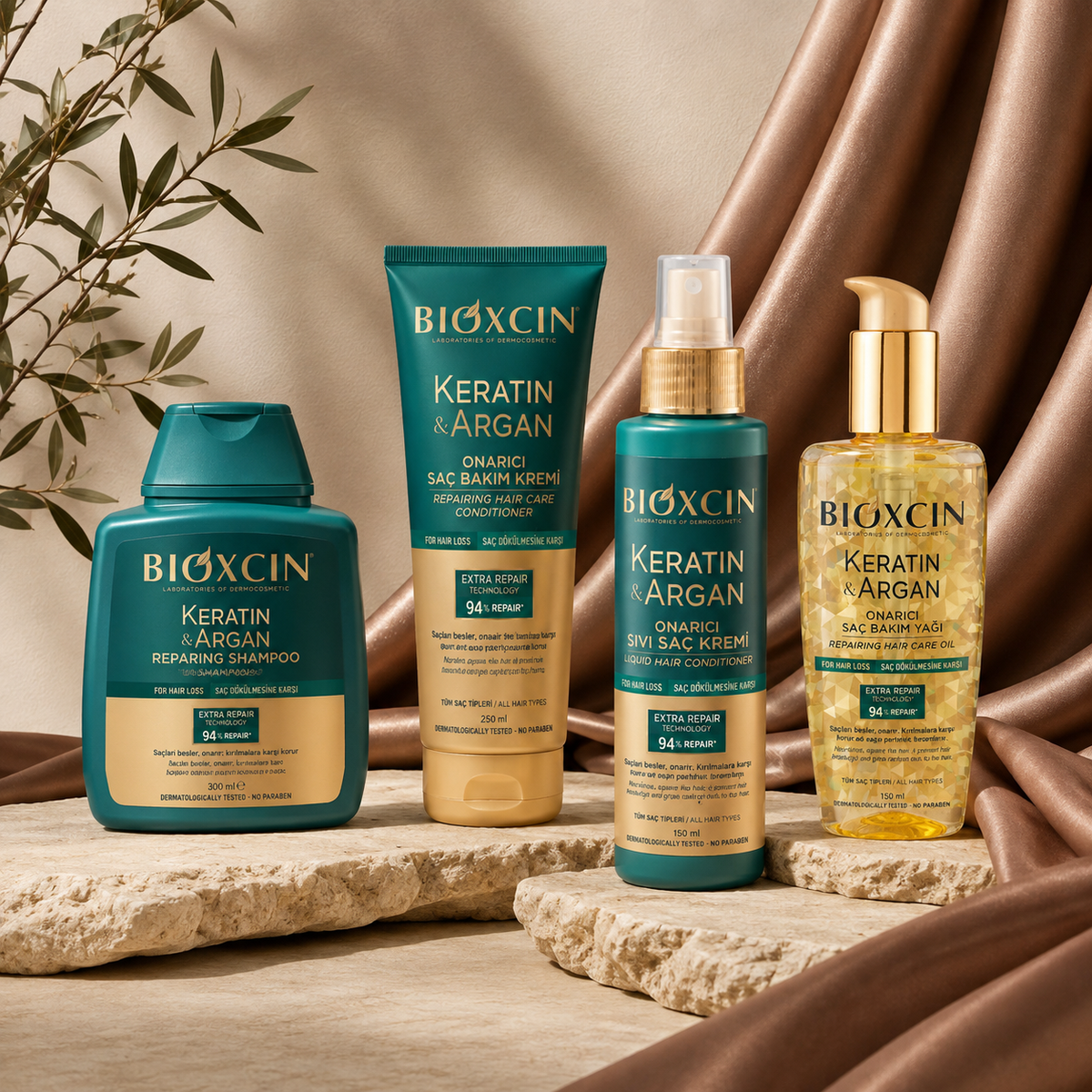 Four BIOXCIN Keratin & Argan haircare products arranged on a stone slab with draped satin fabric and leafy branches in the background: a shampoo bottle, a conditioner tube, a spray bottle, and a treatment cream bottle.